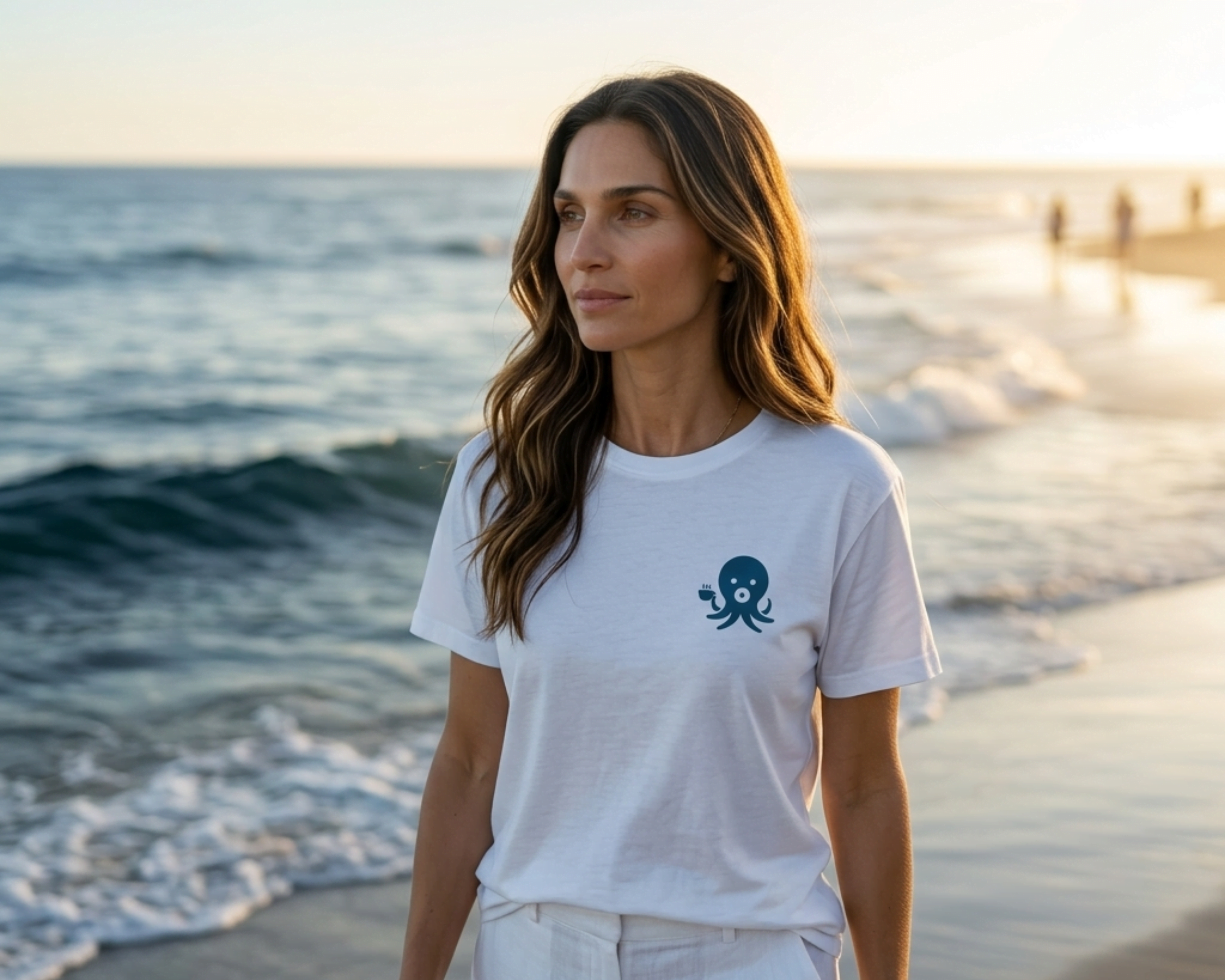 Woman walking on a California beach at golden hour wearing a white Wonder Waves Ollie the Octopus graphic tee and white linen pants