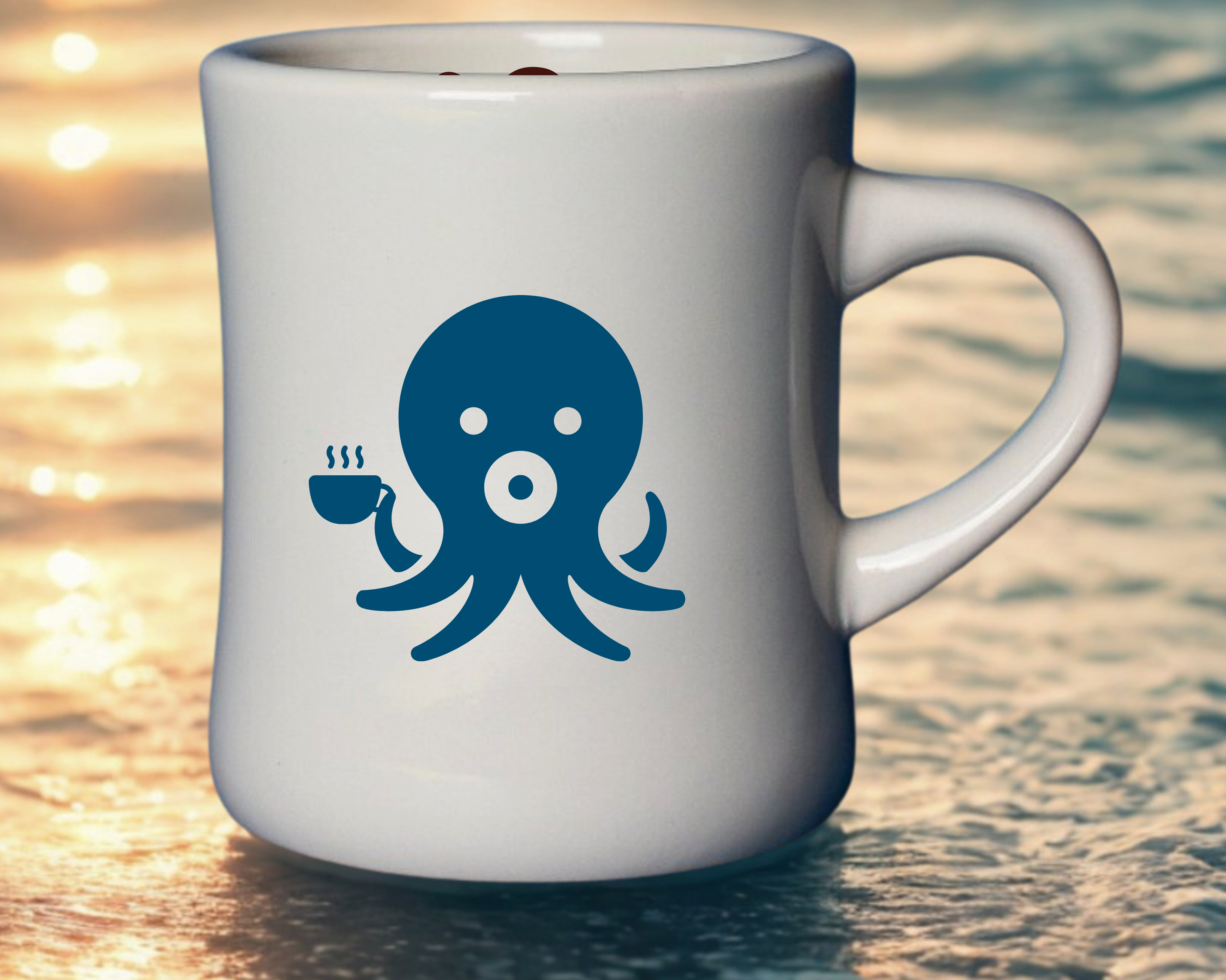 White mug with blue octopus design on a beach with ocean in the background