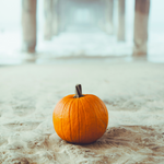 Orange pumpkin resting on a sandy beach beneath a pier on a misty day