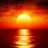 A vivid orange and red sunset over the ocean, with the sun glowing low on the horizon and its golden reflection shimmering across the calm water