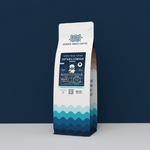 A 12oz bag of Wonder Waves "Captain's Compass" Honduras specialty coffee from La Paz. The white bag features teal wave patterns at the base and a navy blue label with a sea captain illustration. It is positioned against a solid dark navy background, highlighting the premium whole bean coffee.