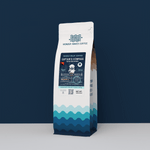 A 12oz bag of Wonder Waves "Captain's Compass" Papua New Guinea specialty coffee from Jiwaka Province. The white bag features teal wave patterns at the base and a navy blue label with a sea captain illustration. It is positioned against a solid dark navy background, highlighting the premium whole bean coffee.