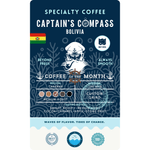 Product label for Wonder Waves "Captain's Compass" Coffee of the Month featuring Bolivia Caranavi District beans. The navy blue label shows a white sea captain illustration and the Rwanda flag. It is a medium roast with bright acidity and tasting notes of cocoa, caramel, apples, and stone fruit, grown at 1,500 MASL.