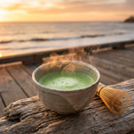 A steaming bowl of Wonder Waves Coffee whisked premium Japanese matcha with a traditional bamboo chasen whisk on a weathered wooden pier. Cinematic golden hour sunset over the ocean in the background.