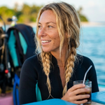A smiling female scuba diver in a black wetsuit with blonde braided hair, enjoying a glass of iced Wonder Waves Coffee Diver's Delight cold brew specialty coffee on a boat after a dive. The tropical blue ocean and diving gear are visible in the background, highlighting the refreshing lifestyle of specialty coffee for ocean adventurers.