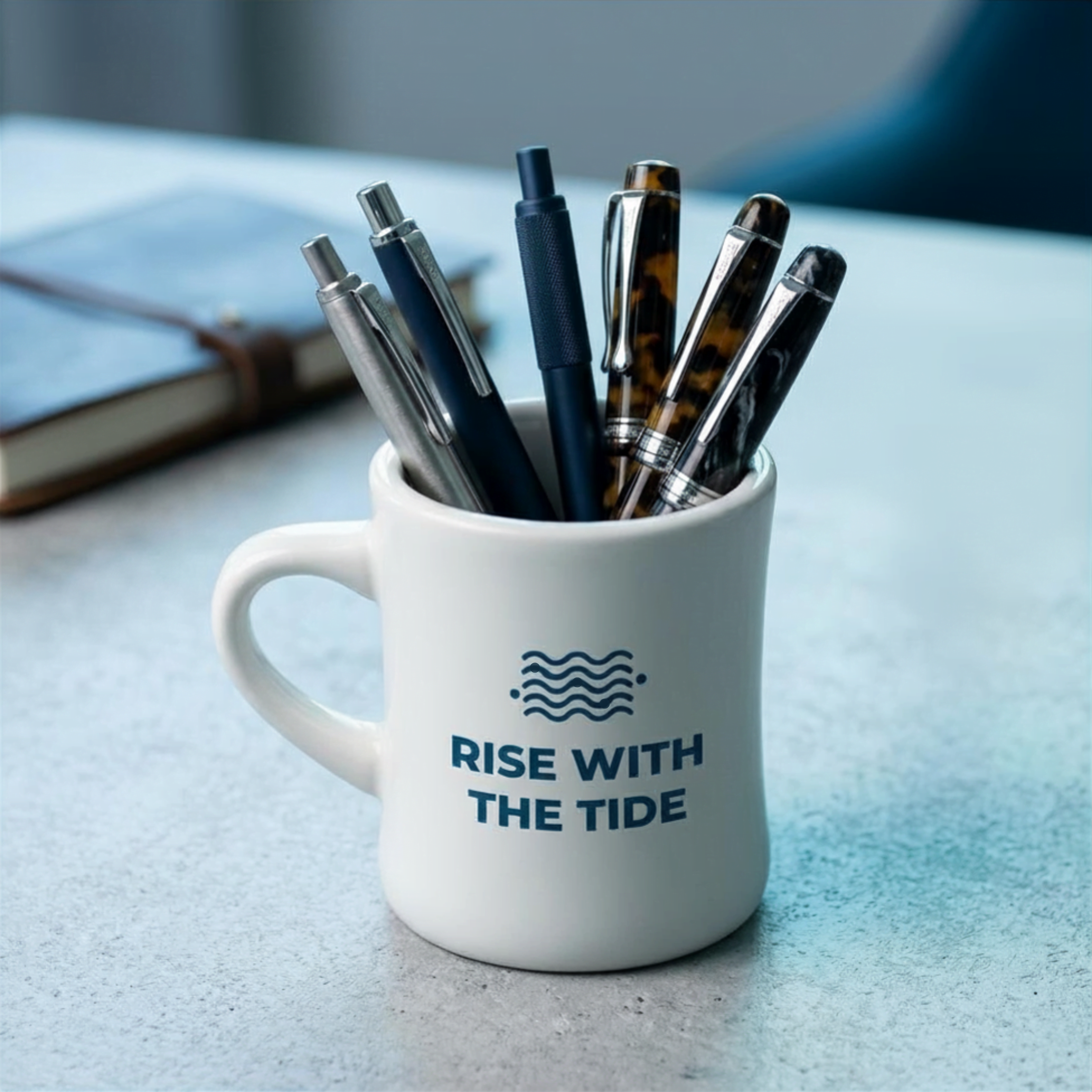Close-up of a white Wonder Waves Coffee "Rise With The Tide" mug filled with pens and pencils on a textured grey studio desk next to a leather-bound journal.