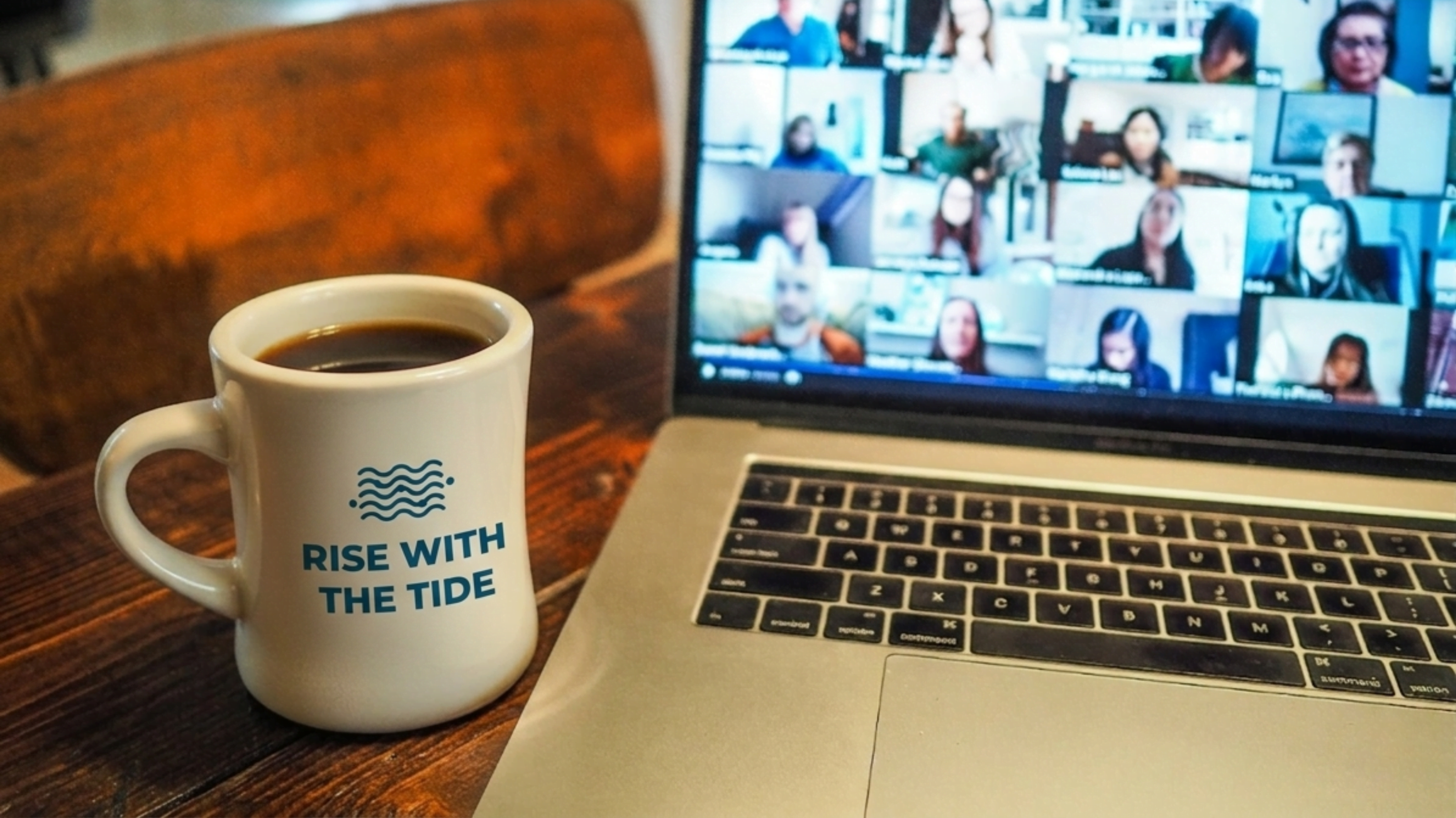 A Wonder Waves Coffee "Rise With The Tide" branded diner mug sits on a wooden desk next to a laptop displaying a virtual team meeting with several participants on screen.