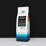 A 12oz bag of Wonder Waves Marea Magnifica Italian Roast. The white bag features a dark blue wave pattern and a black label with a white wave icon and the Italian flag. Set against a minimalist black and white background, the label highlights a deep, elegant, and intense flavor profile.
