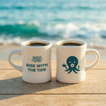 Two Wonder Waves Coffee white ceramic diner mugs filled with black coffee on a weathered wooden surface at the beach showing both the Rise With The Tide wave design and Ollie the Octopus sides with ocean waves in the background